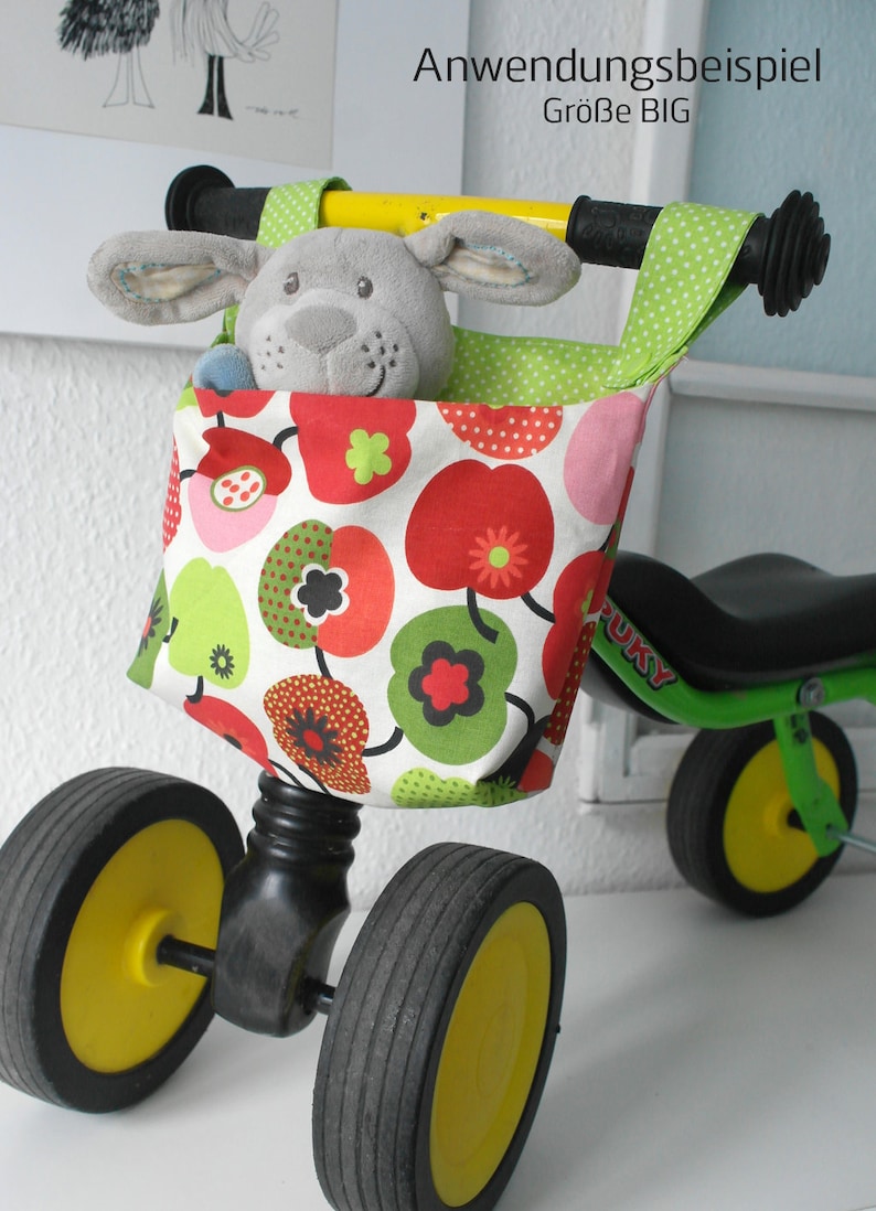 May include: A green and black tricycle with yellow wheels and a fabric storage bag with a red, green and white floral pattern. The bag is attached to the handlebars and contains a grey stuffed animal.