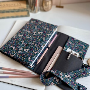 Könnte beinhalten: Ein offenes Notizbuch mit einem floral gemusterten Einband in Teal, Rosa und Weiß. Im Inneren sind ein Bleistift, kleine Notizbücher und ein Stift zu sehen. Ein passendes Blumenband mit Knopfverschluss sichert das Notizbuch. Mehrere Bleistifte liegen auf den offenen Seiten.