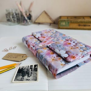 Könnte beinhalten: Ein Notizbuch mit floralem Muster und Knopfverschluss, mit rosa, weißen und lila Blumen. Das Notizbuch liegt auf einer linierten Seite, zusammen mit einem Vintage-Foto, Bleistiften und einem Etikett mit der Aufschrift "handmade with love".