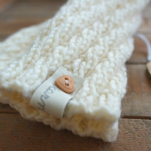 Könnte beinhalten: Ein weißes gestricktes Stirnband mit einem braunen herzförmigen Knopf und einem Etikett mit der Aufschrift "Kourt".