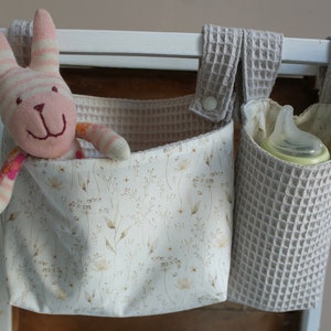 May include: Two fabric hanging organisers with a white background and a floral pattern. One organiser has a pink and white striped stuffed bunny inside. The other organiser has a clear baby bottle inside. Both organisers have a grey waffle-textured fabric exterior.