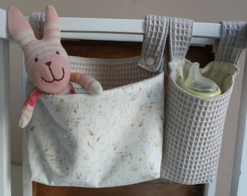 May include: Two fabric hanging organisers with a white background and a floral pattern. One organiser has a pink and white striped stuffed bunny inside. The other organiser has a clear baby bottle inside. Both organisers have a grey waffle-textured fabric exterior.