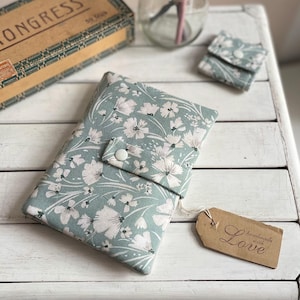 Könnte beinhalten: Stoffetui in Türkis mit weißem Blumenmuster und Knopfverschluss. Ein kleines passendes Täschchen und ein Anhänger mit der Aufschrift "handmade with Love" sind ebenfalls zu sehen. Das Etui ist wahrscheinlich für ein Telefon oder ein kleines Notizbuch.