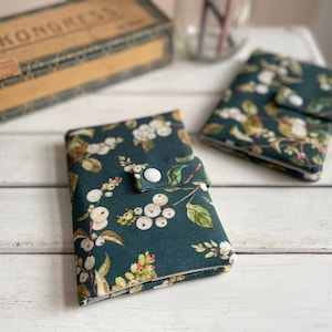 Könnte beinhalten: Zwei stoffbezogene Notizbücher mit einem dunkelblauen Blumenmuster mit weißen Beeren und grünen Blättern. Jedes Notizbuch hat einen weißen Knopfverschluss. Die Notizbücher liegen auf einer weißen Holzoberfläche.