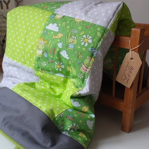 Könnte beinhalten: Eine Patchwork-Babydecke mit grünen, weißen und grauen Paneelen. Die grünen Paneele zeigen ein skurriles Frosch- und Blumenmuster. Die Decke liegt auf einem kleinen Holzbett, mit einem Anhänger mit der Aufschrift "Love".