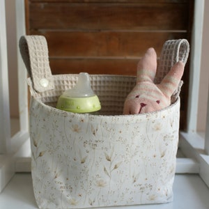 May include: A white fabric storage bin with a floral pattern and two beige fabric handles. The bin contains a green baby bottle and a pink and white striped stuffed bunny.