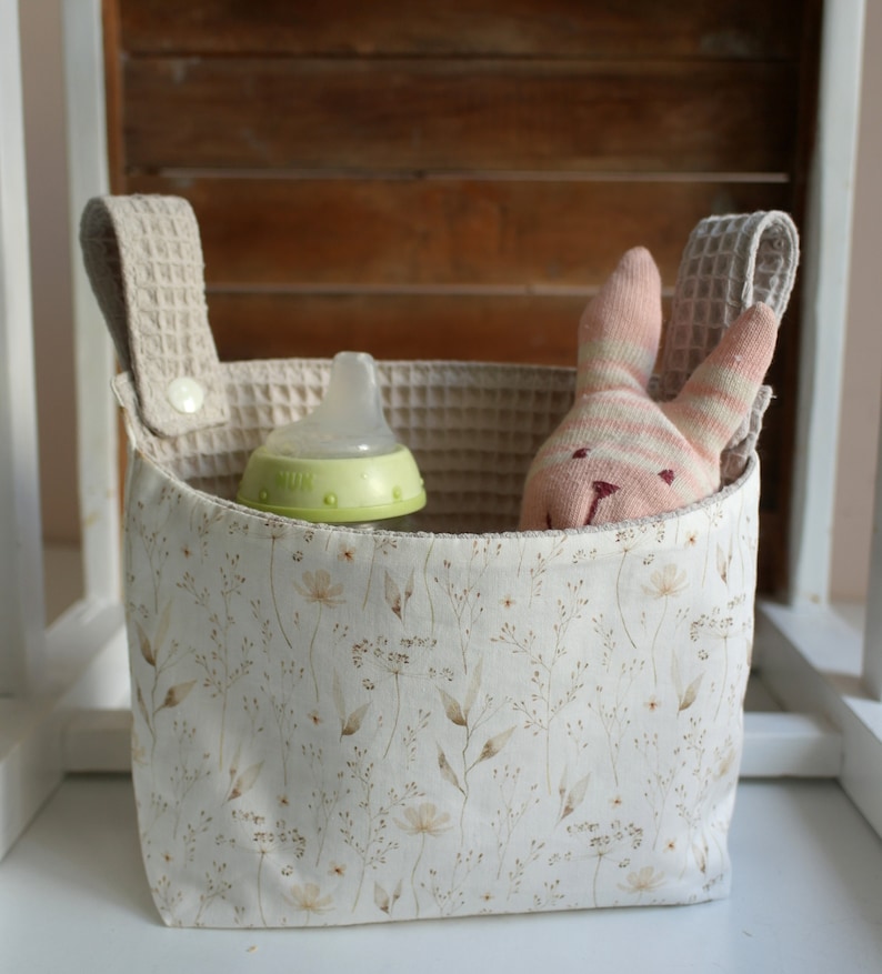 May include: A white fabric storage bin with a floral pattern and two beige fabric handles. The bin contains a green baby bottle and a pink and white striped stuffed bunny.