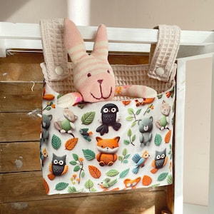 May include: A hanging storage bag with a woodland animal theme. The bag is white with colorful forest creature appliqués. A pink and white striped bunny toy is inside the bag. The bag is attached to a wooden frame with a beige fabric strap.
