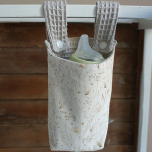 May include: A beige fabric bottle holder with a floral pattern and two beige fabric straps with white buttons. A clear baby bottle is inside the holder.