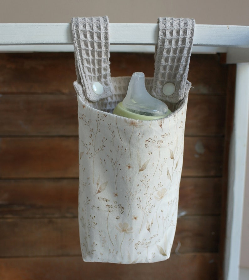 May include: A beige fabric bottle holder with a floral pattern and two beige fabric straps with white buttons. A clear baby bottle is inside the holder.