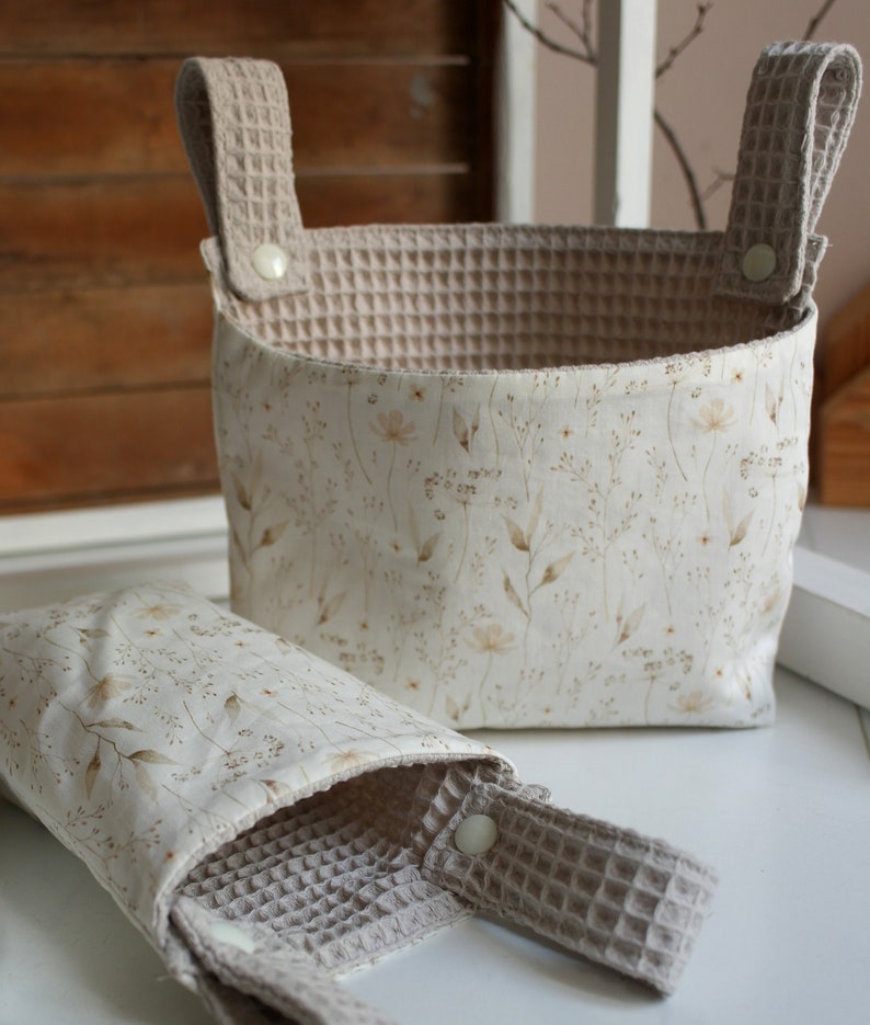 May include: Two beige fabric storage baskets with white floral patterns and waffle-textured handles. The larger basket is taller and wider than the smaller basket.