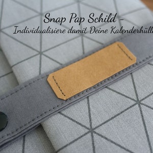 May include: A gray fabric calendar cover with a geometric pattern and a brown leather strap with a snap closure. The strap has a label that says "Snap Pap Schild - Individualisiere damit Deine Kalenderh&uuml;lle".