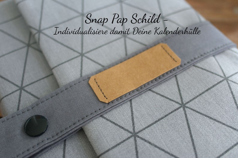 May include: A gray fabric calendar cover with a geometric pattern and a brown leather strap with a snap closure. The strap has a label that says "Snap Pap Schild - Individualisiere damit Deine Kalenderh&uuml;lle".