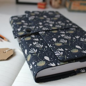 Könnte beinhalten: Ein dunkelblauer Notizbuchumschlag mit einem weißen Blumenmuster. Der Umschlag hat eine Klappe, die sich über die Vorderseite des Notizbuchs klappen lässt. Die Klappe hat einen Knopfverschluss. Das Notizbuch liegt auf einer weißen Oberfläche.
