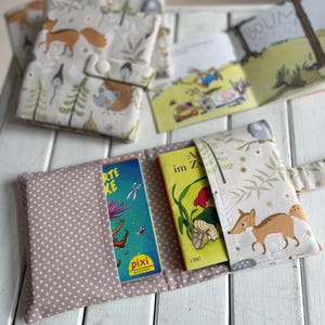Könnte beinhalten: Eine handgefertigte Buchhülle und passende Büchertaschen. Die Hülle ist hellrosa mit weißen Punkten. Die Taschen und die Umschläge der Hülle zeigen einen Waldtierdruck mit Füchsen, Igeln und Bäumen. Zwei kleine Bücher sind sichtbar.