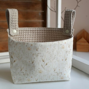 May include: A beige fabric storage basket with two handles. The basket has a white floral pattern and a beige waffle-textured lining.