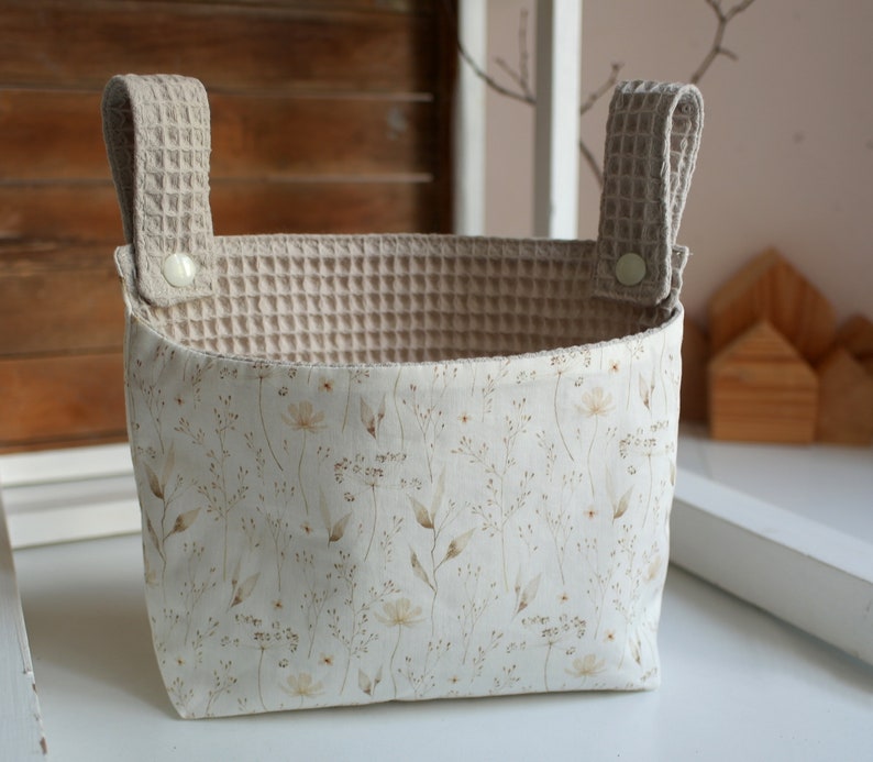 May include: A beige fabric storage basket with two handles. The basket has a white floral pattern and a beige waffle-textured lining.