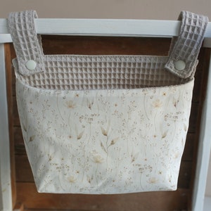 May include: A beige fabric hanging organiser with a floral pattern and waffle-textured straps. The organiser is hanging on a white wooden bar.
