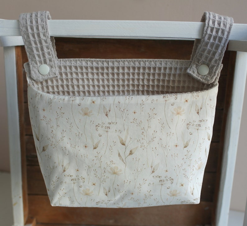 May include: A beige fabric hanging organiser with a floral pattern and waffle-textured straps. The organiser is hanging on a white wooden bar.