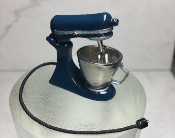 以下が含まれることがあります： 銀色のボウルと黒いコードが付いたミニチュアの青いスタンドミキサー。ミキサーは透明な台座の上に置かれています。