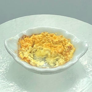 May include: A small white ceramic dish with a scalloped rim filled with a golden yellow casserole. The casserole is topped with a crispy, golden brown crust.