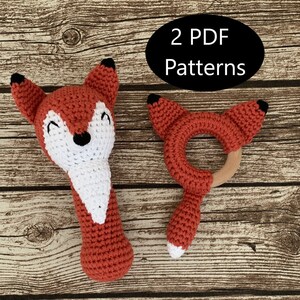 May include: Two crochet fox toys, one is a rattle and the other is a teether. The rattle is red with white accents and black eyes. The teether is red with a wooden ring in the center. The toys are on a wooden background. 2 PDF Patterns.