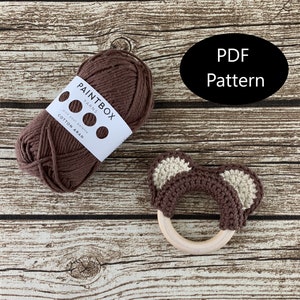 Peut inclure: Un hochet en forme d'ours en crochet marron avec des oreilles beiges attachées à un anneau en bois. La laine utilisée est de la laine d'aran en coton marron de Paintbox Yarns.