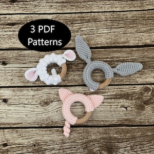 Op de afbeelding: Drie gehaakte baby-bijtringen in de vorm van een lam, een konijn en een varken. Elke bijtring is bevestigd aan een houten ring. De bijtringen liggen op een houten achtergrond. 3 PDF-patronen.