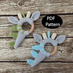 May include: Two crocheted baby teethers in the shape of a dinosaur with wooden rings. One teether is grey with green accents and the other is grey with blue accents. The text "PDF Pattern" is visible in the image.
