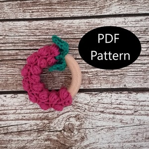 May include: A pink crocheted teething ring with green leaves attached to a wooden ring. The ring is on a wooden background. PDF Pattern.