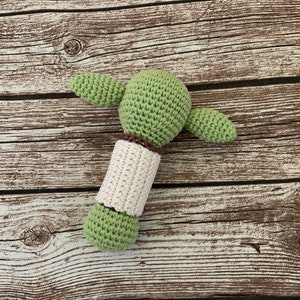 K&ouml;nnte beinhalten: Gr&uuml;nes H&auml;kel-Rassel f&uuml;r Babys in Form einer Figur mit braunen Garn-Akzenten.