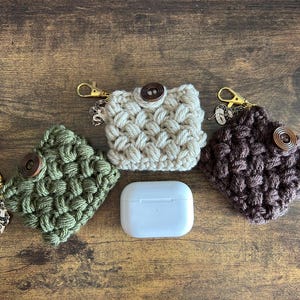 May include: Three square crochet pouches in olive green, cream, and dark brown, each featuring a dark wooden button closure and a gold-toned metal lobster clasp with a small silver-toned snake charm. A white wireless earbud case sits in the centre.