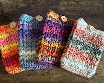 May include: Four colorful, handmade crochet cozies in shades of orange, pink, purple, blue, and gray. Each cozy features a textured stitch pattern and is secured with a wooden button. These cozies are designed to fit a standard-sized coffee cup.
