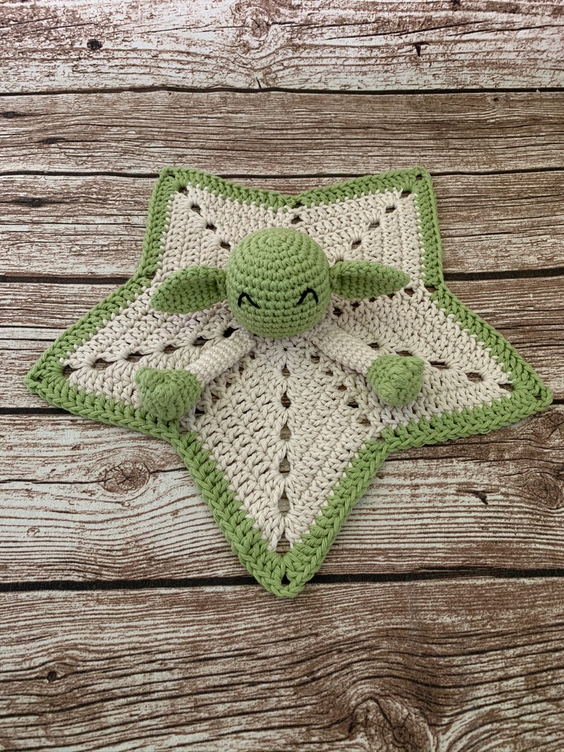 K&ouml;nnte beinhalten: Eine gr&uuml;ne und wei&szlig;e geh&auml;kelte Sternen-Babydecke mit einer gr&uuml;nen geh&auml;kelten Figur in der Mitte. Die Figur hat einen runden Kopf mit schwarzen Augen und einem kleinen Mund.