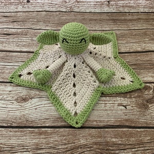 K&ouml;nnte beinhalten: Eine gr&uuml;ne geh&auml;kelte Babydecke mit einer geh&auml;kelten Yoda-Figur in der Mitte. Die Decke ist wei&szlig; mit einem gr&uuml;nen Rand.