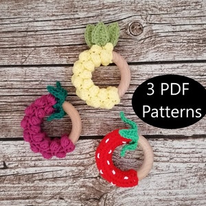 May include: Three crochet teething rings in the shapes of a pineapple, strawberry, and raspberry. The rings are attached to wooden rings. The text "3 PDF Patterns" is written on a black oval in the center of the image.