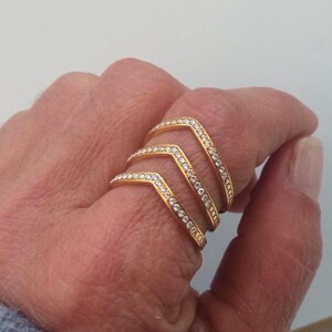 May include: A gold-coloured ring featuring three chevron-shaped bands, each set with small, clear gemstones. The ring is worn on a finger, highlighting its design and sparkle. The ring's design is contemporary and stylish.