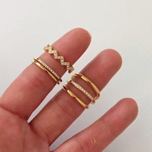 Peut inclure: Gros plan de plusieurs bagues dorées. Une bague a un motif en zigzag avec de petites pierres transparentes. Deux bagues sont des anneaux simples et deux ont une rangée de petites pierres transparentes. Les bagues sont sur un doigt sur fond blanc.