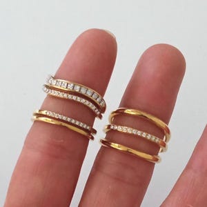 Può includere: Due dita adornate da anelli d'oro. Un dito mostra tre anelli: due fasce lisce e una con piccole pietre trasparenti. L'altro dito ha tre anelli: uno con pietre quadrate e due con piccole pietre trasparenti.