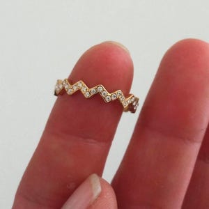 Könnte beinhalten: Ein goldfarbener Ring mit Zickzack-Muster, besetzt mit kleinen, klaren Edelsteinen. Der Ring wird zwischen zwei Fingern gehalten, was sein filigranes und aufwendiges Muster zeigt. Das Design des Rings ist eine sich wiederholende Reihe von Chevron-Formen.