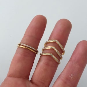 Puede incluir: Dos anillos dorados con piedras claras y brillantes. Uno es una banda simple con una fila de piedras. El otro es un conjunto de tres anillos en forma de cheurón, también adornados con piedras. Los anillos se muestran en dedos.