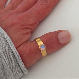 Op de afbeelding: Een goudkleurige ring met een ronde, lichtblauwe en roze edelsteen. De ring heeft een eenvoudig bandontwerp met een bezel-zetting voor de edelsteen. De ring zit aan een duim.