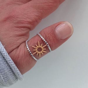 Op de afbeelding: Een zilveren ring met een gouden zonnemotief. De ring heeft twee banden, één boven en één onder het zonnemotief. Het zonnemotief is gecentreerd op de vinger, wat een uniek en opvallend accessoire creëert.