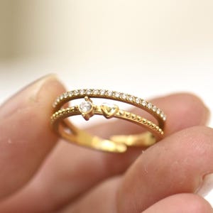 Peut inclure: Une bague dorée à deux rangs. La rangée supérieure est sertie de petites pierres transparentes. La rangée inférieure présente une petite pierre transparente et un motif en forme de cœur. La bague est tenue dans une main.