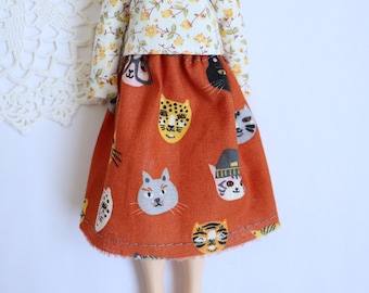 Puede incluir: Un conjunto de muñeca que consta de una parte superior crema con un estampado floral amarillo y una falda color óxido. La falda está decorada con varias caras de gato. El conjunto es un diseño divertido y juguetón.