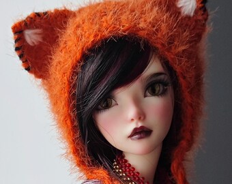 Peut inclure: Une poupée aux longs cheveux noirs portant un chapeau orange duveteux avec des accents blancs. La poupée a un tatouage floral sur le bras et porte un haut violet clair avec un collier argenté.