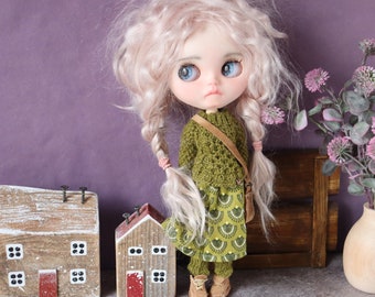 Puede incluir: Una muñeca con el pelo rubio rizado que lleva un jersey verde y una falda verde y blanca. La muñeca está de pie en un estante con dos pequeñas casas de madera y un jarrón de flores.