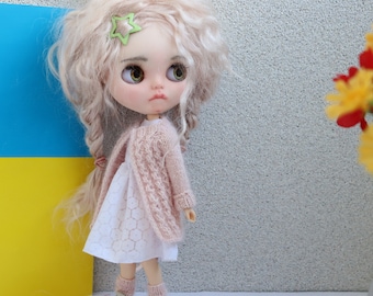 May include: A doll with long, blonde, curly hair wearing a pink knitted cardigan over a white dress. The doll has green eyes and is looking to the right. The doll is standing in front of a blue and yellow background.