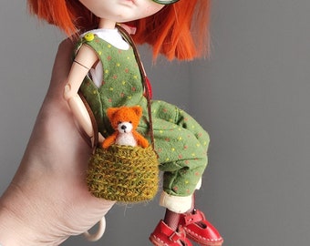 May include: A doll with red hair, green-framed glasses, and a green jumpsuit with yellow star patterns. The doll has a small brown teddy bear in a green knitted bag and wears red shoes. The doll's hair is styled with small yellow and orange flowers.