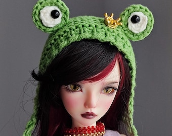 Peut inclure: Une poupée aux cheveux noirs et rouges porte un chapeau de grenouille vert crocheté avec une couronne dorée. La poupée porte un collier de perles rouges et un tatouage sur le bras. La poupée porte un débardeur lavande.